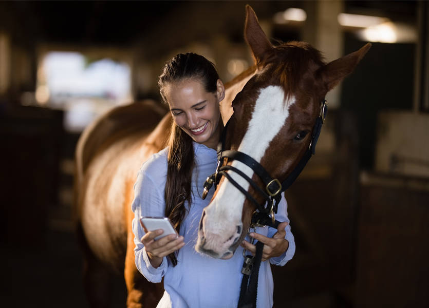 woman looking at phone with horse looking over her shoulder