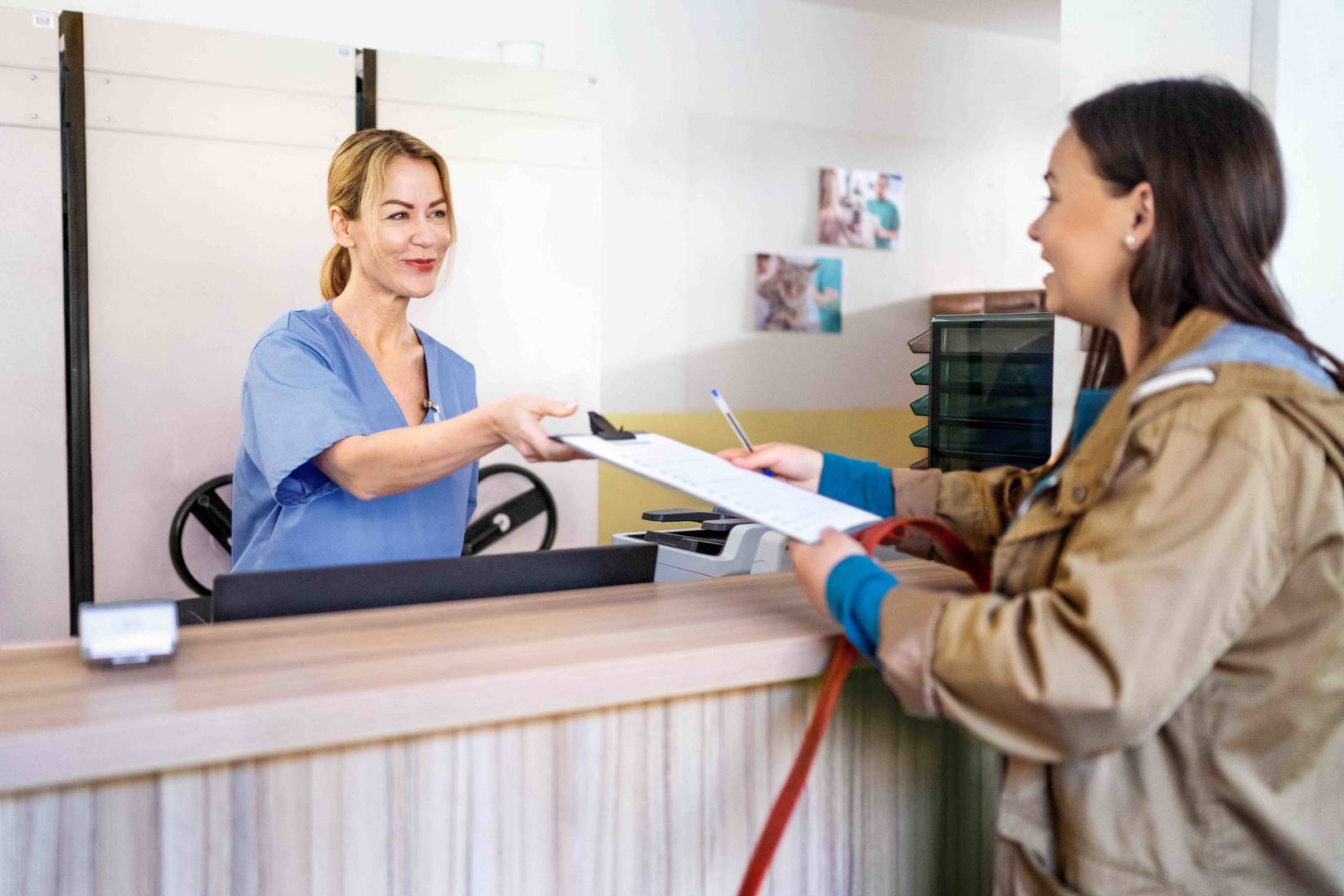 A smiling woman in blue scrubs hands documents to a pet owner at a veterinarian's reception desk.